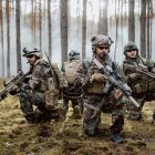 Four fully equipped, middle-aged soldiers in camouflage uniforms form a line, ready to fire, aiming with their rifles. A military operation in action, a unit standing in a dense forest.